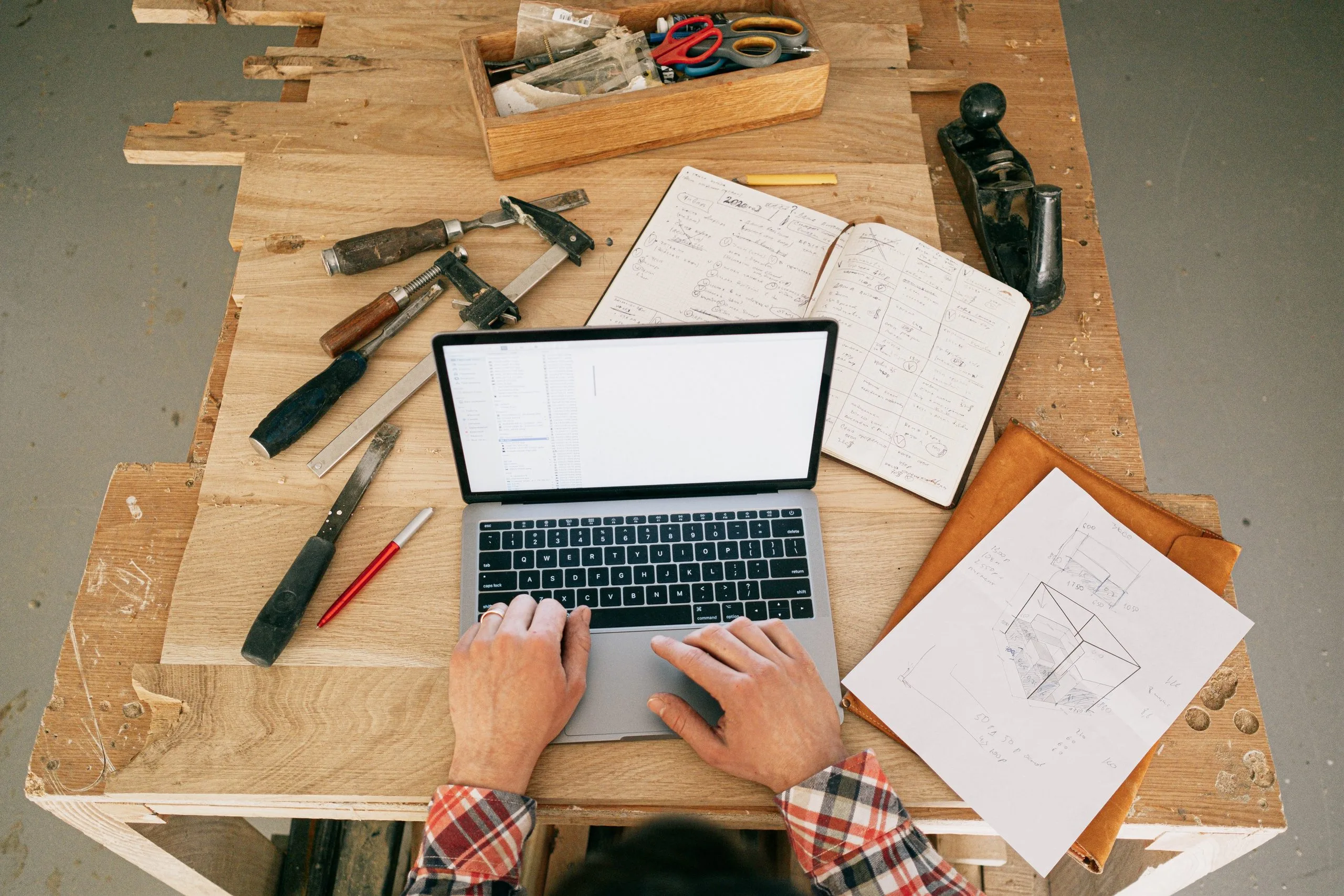 Planungssituation mit Laptop, Werkzeugen und handschriftlichen Skizzen auf einer Holzwerkbank.