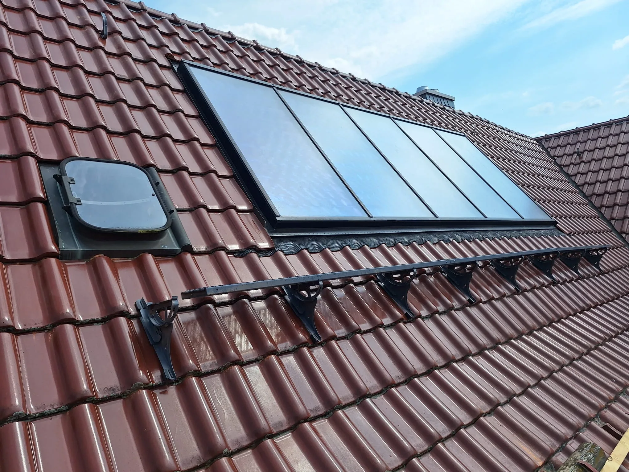 Solarthermie-Kollektoren frisch auf einem Ziegeldach mit braunen Tondachpfannen montiert, daneben ein Dachfenster.