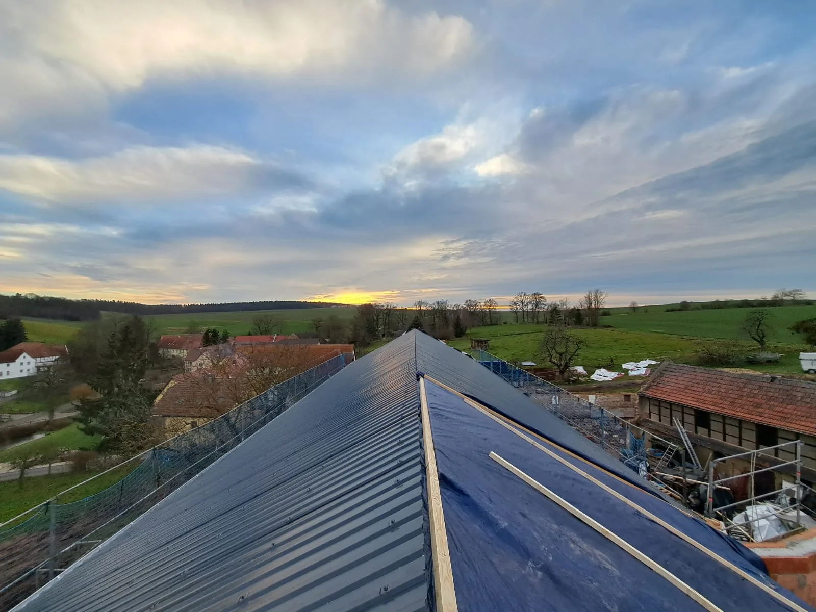 Neu gedeckter Dachstuhl einer Bauernhofsanierung mit dunkler Metalleindeckung im Abendlicht über Feldlandschaft.